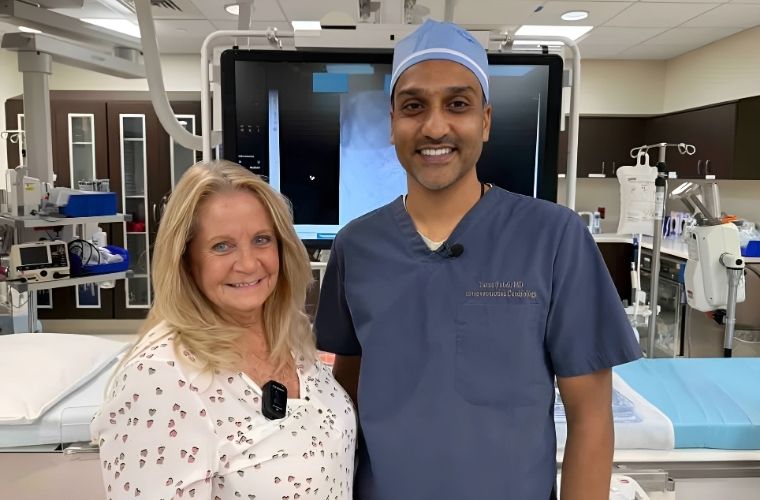 A smiling patient on the left in a white blouse stands with her cardiologist in his blue scrubs in the Cath Lab in front of a monitor.