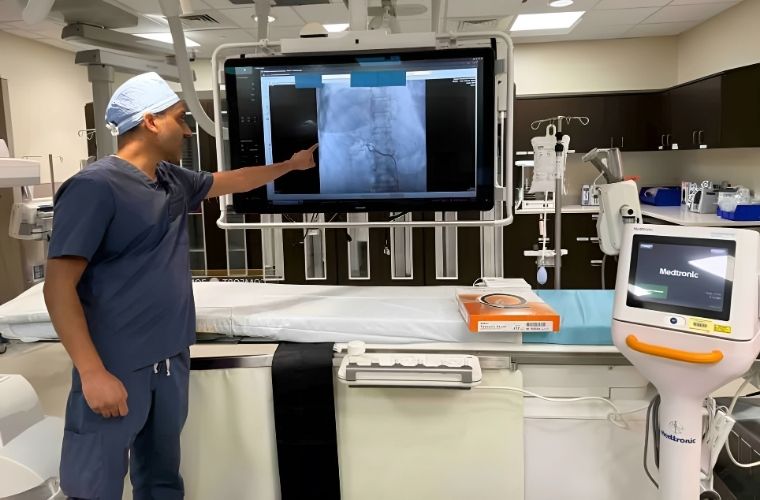 An interventional cardiologist in blue scrubs looks at the large monitor with the image of an angiogram in the Cath Lab.
