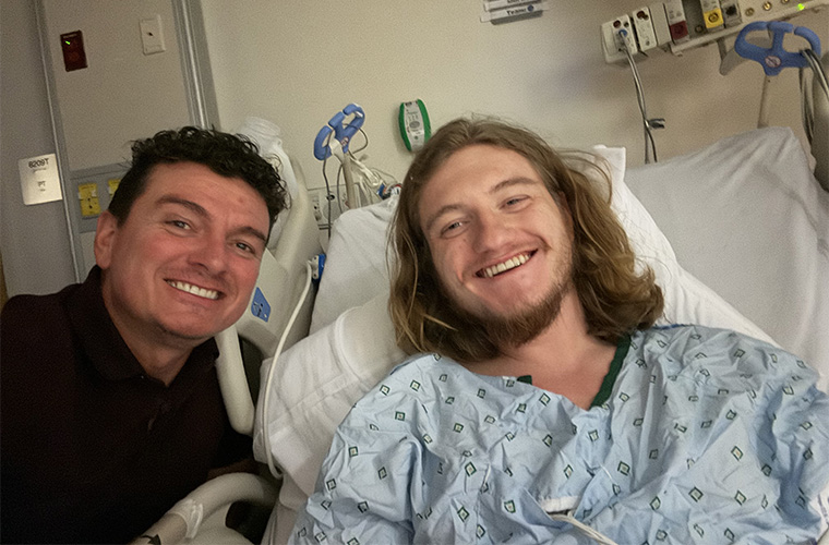 A man with dark hair in a black shirt sits next to a younger man with long hair in a hospital bed.