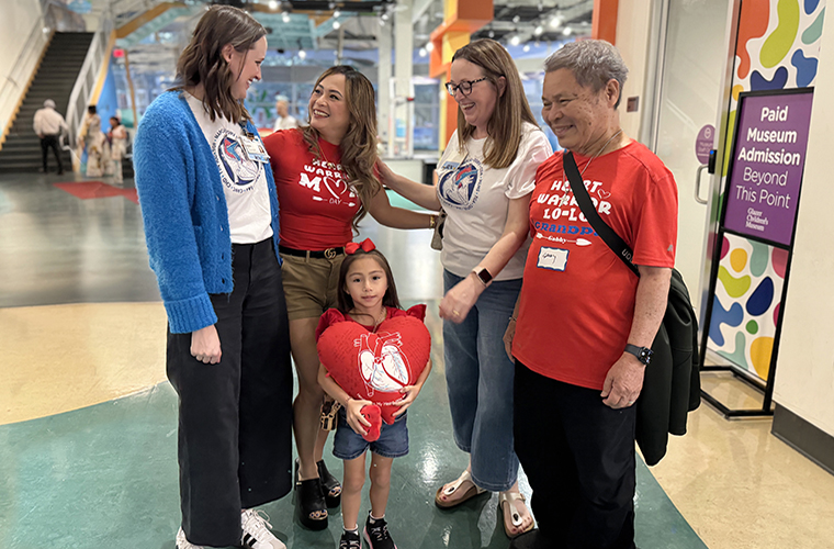 Three women and a man with a little girl, all engaged in conversation and sharing a joyful moment. The little girl is holding a red heart-shaped pillow.