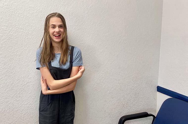 A teen girl smiling with arms crossed, standing in front of a light gray wall next to a blue chair.