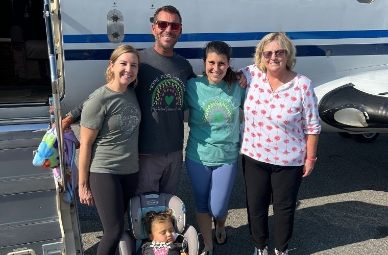 Group photo of four adults standing in front of a private jet, with a young hospital patient in a car seat positioned in the foreground. Two of the adults – the child’s parents - wear matching T-shirts with the text 'Hope for Harper' and a colorful rainbow design. The jet's doorway is open, and the group is standing on an airport tarmac having just returned from Canada where the child received an experimental treatment.