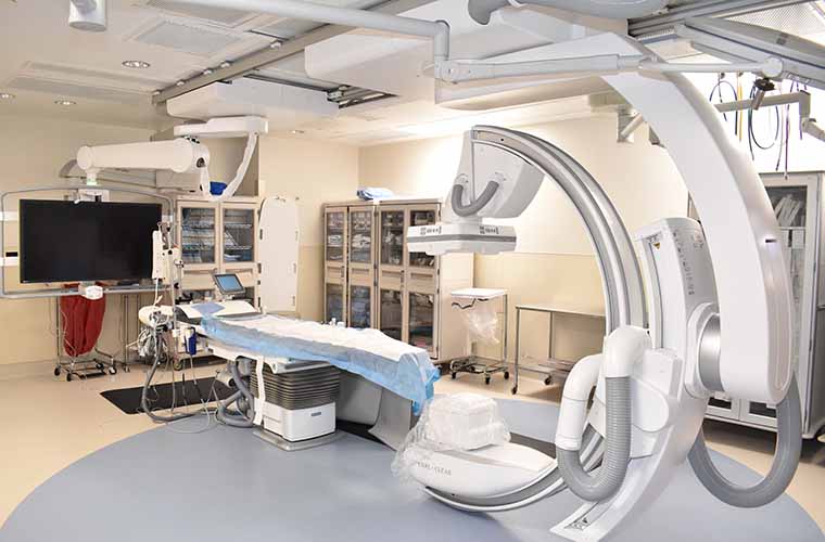 Interior of a modern medical interventional radiology room with a state-of-the-art angiography system and various medical equipment, including monitors and surgical lights.