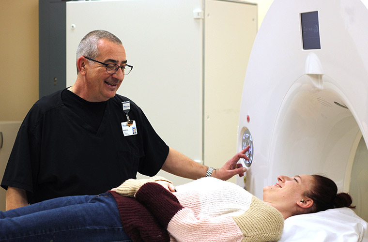 A person wearing black scrubs smiles while looking at another person lying on a medical exam table. The head of the person lying on the table appears to be entering a CT scanning unit.