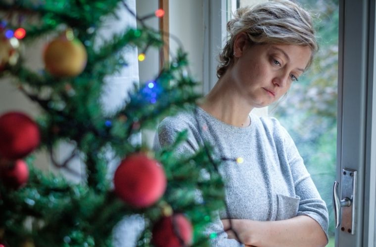 A woman looking down sadly poses next to a festive Christmas tree adorned with lights and ornaments.