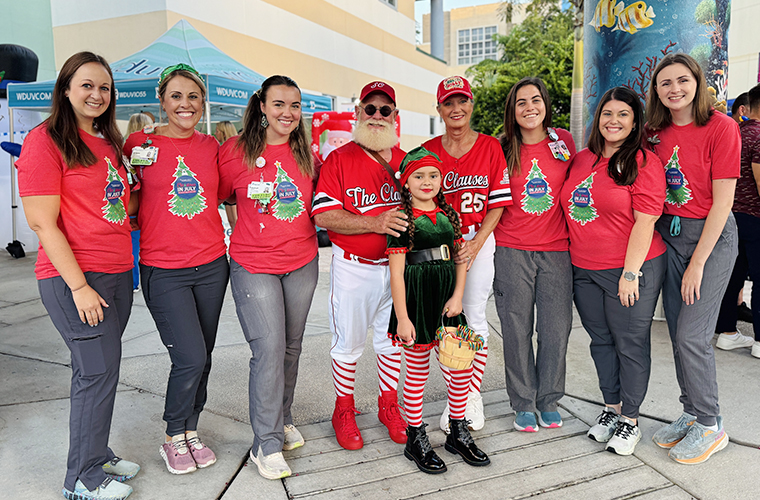A group of people stand together outdoors, with most wearing red Christmas-themed shirts; two are dressed as Santa and Mrs. Claus, and one child is dressed as an elf.