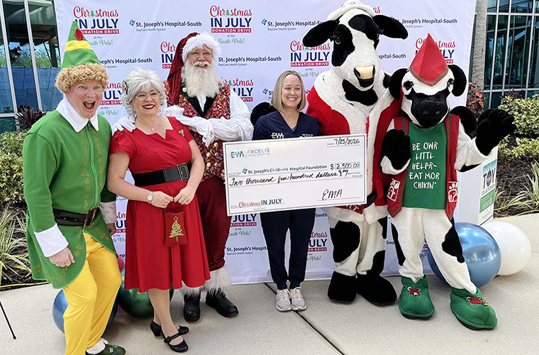 A group of people wearing festive clothing, including costumes and mascots, are standing outdoors and smiling while posing with a large check.