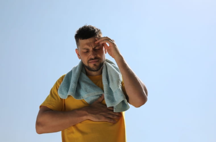 A man in a yellow shirt with a blue towel around his neck is holding his head and chest, expressing stress and fatigue from the high temperatures.