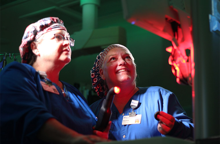 Two women in surgical caps are looking at a monitor in a medical setting. One is holding a probe with a red light.