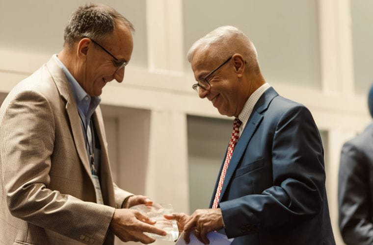 Two professionally dressed men smile as they accept a clear award plaque during a recognition event. One wears a beige blazer and glasses, the other a navy suit with a red patterned tie.
