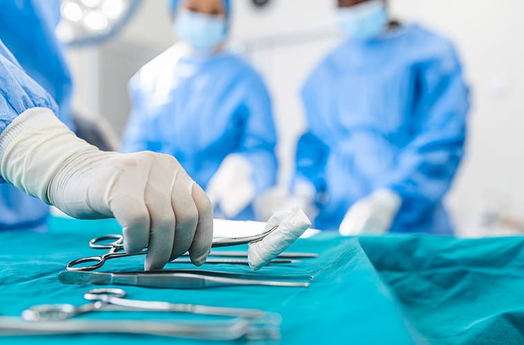 Gloved hand taking surgical instrument off table with a group of surgeons standing in background of operating room.