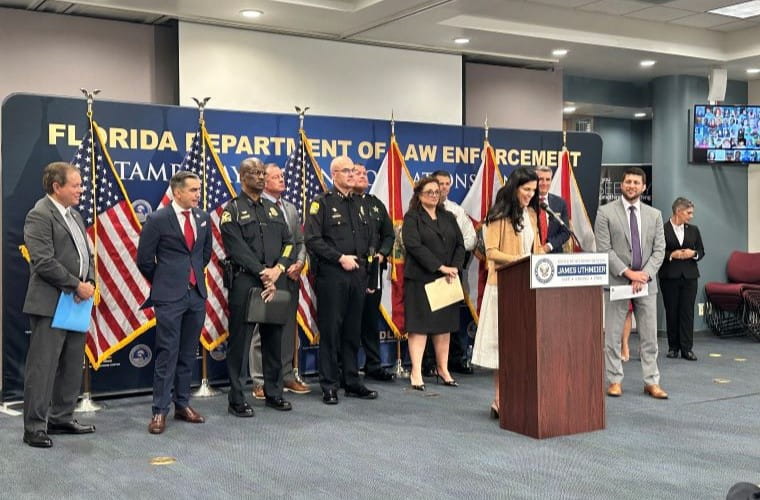 A group of individuals, including law enforcement officers and civilians, standing at a press conference. One person is speaking at a podium with a sign reading "Department of Law Enforcement." The background displays several American flags and the podium bears the seal of Florida Attorney General James Uthmeier.