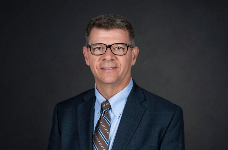 A smiling headshot of C. Todd Jones. He is wearing glasses with a dark navy suit jacket, a light blue shirt with a multi-striped tie.