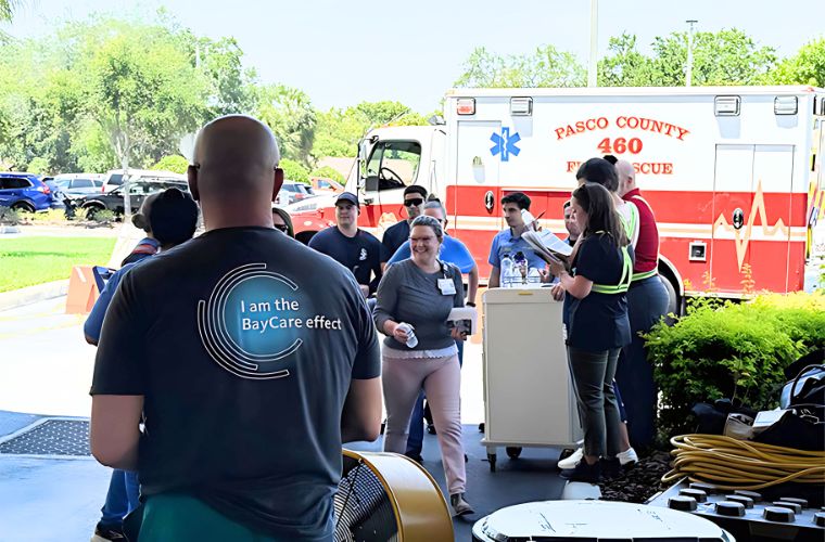 A group of people participating in an outdoor event near a Pasco County Fire Rescue ambulance. Some are standing around a table with refreshments, and one person is addressing others near a large fan.
