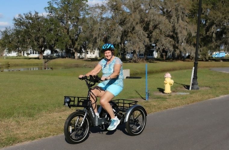 A woman wearing blue shorts and top with a matching blue bike helmet smiles as she rides her electric bike down the road.