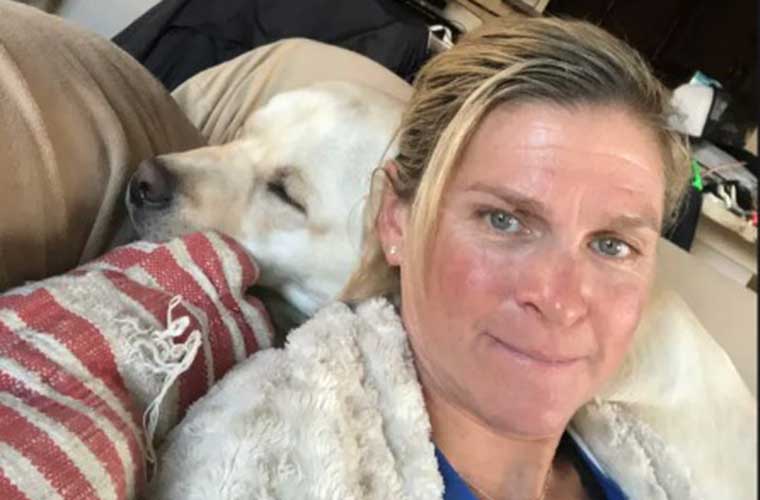 A woman wrapped in a blanket sits on a couch with a dog resting on the back of the couch.