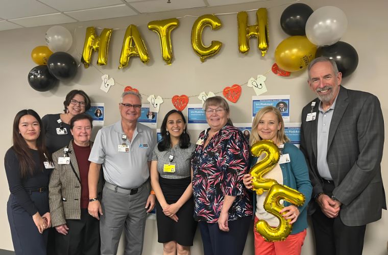 A group of smiling people dressed in street clothes stand underneath a banner of balloons and ballon letters to celebrate an event.