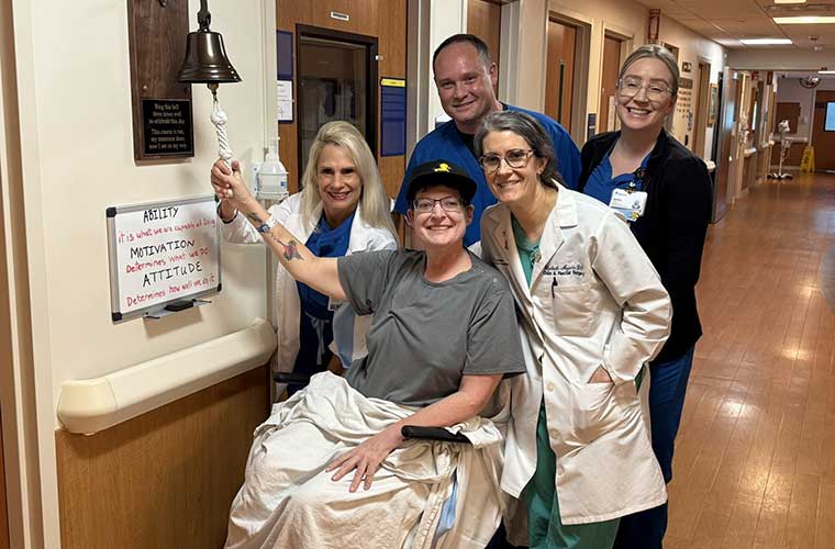 A woman sitting in a chair and wearing a baseball cap, T-shirt and glasses prepares to ring a bell while surrounded by three women and a man who are all dressed in medical scrubs.