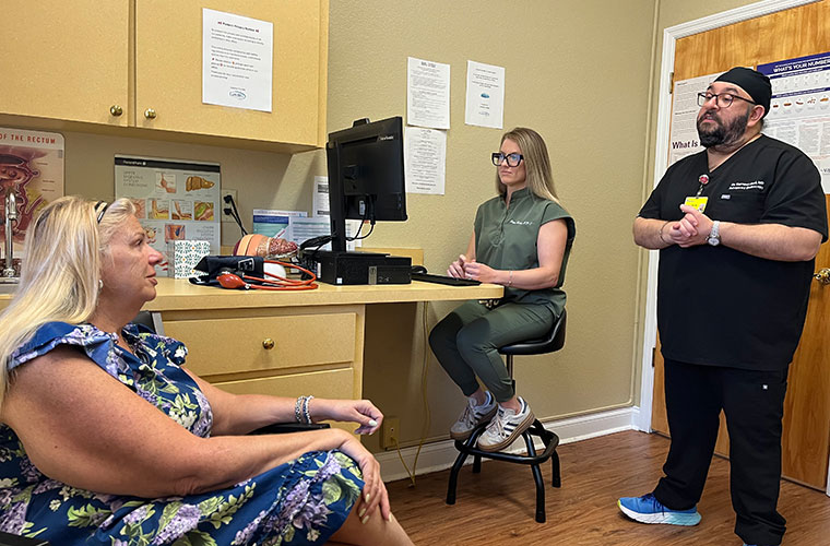 A woman with blond hair wearing a flower print dress who is sitting in a chair talks with a man wearing black hospital scrubs as another woman wearing scrubs works on a computer. 