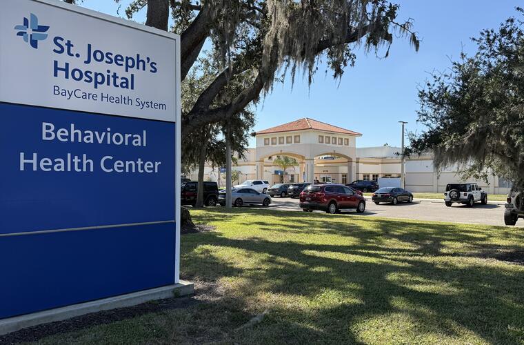 A large roadside sign for St. Joseph’s Hospital’s Behavioral Health Center in Tampa, Florida is seen in front of the facility building, with cars parked and trees providing shade.