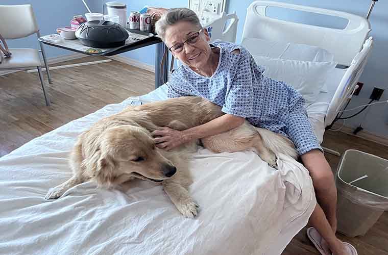 Person in a hospital bed petting a golden retriever, with a bedside table holding personal items nearby. 