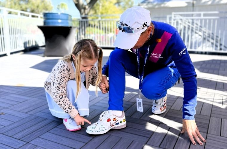 A young girl kneels beside a golf professional, and while holding hands, points to her hand-drawn designs on a pair of custom white sneakers during The Annika.