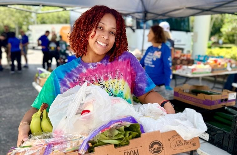  A smiling woman in a colorful tie-dye shirt holds a tray of fresh produce, including bananas and leafy greens, at a food distribution tent during the BayCare Kids Family Expo.