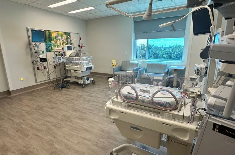 Interior of a modern hospital neonatal intensive care unit featuring an incubator, medical monitors, and a recliner, with a calming artwork hanging on the wall and a view of trees through the window.