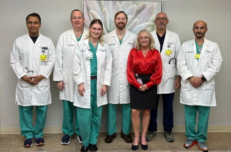 The multi-disciplinary Heart and Valve team stands proud with the six physicians in white lab coats and one woman in red standing in a conference room setting. 