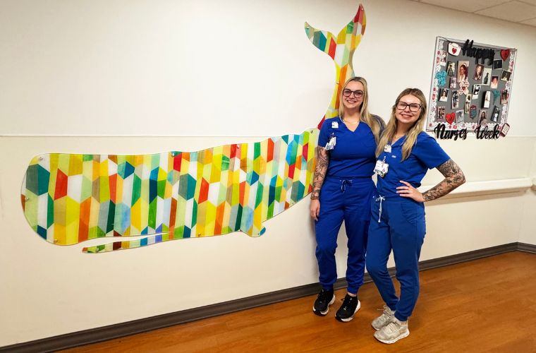 Two women in blue medical scrubs stand in a hallway beside a colorful geometric whale mural, with a “Happy Nurses Week” board on the wall.