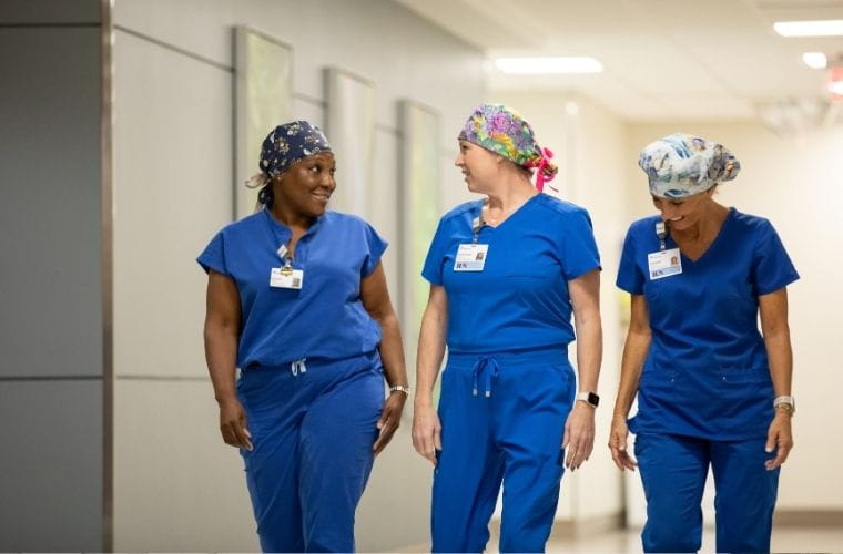 Three women dressed in blue scrubs walk side-by-side down a hospital hallway, smiling and engaged in conversation.