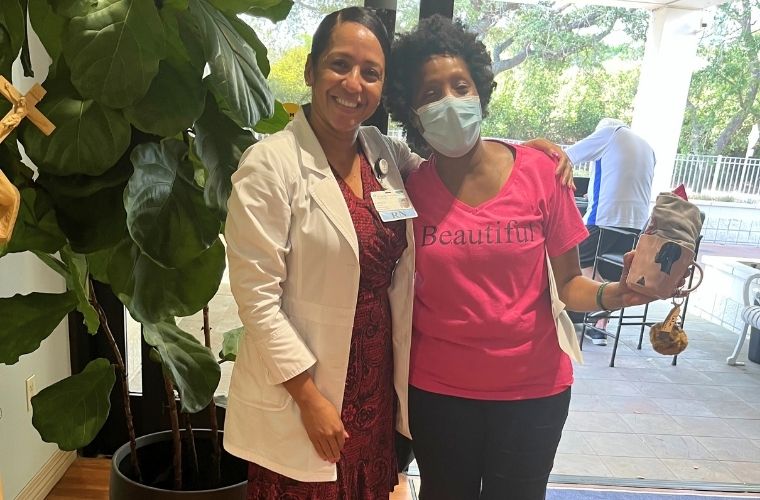 A nurse navigator smiles alongside a patient wearing a mask, standing together indoors near a large plant.