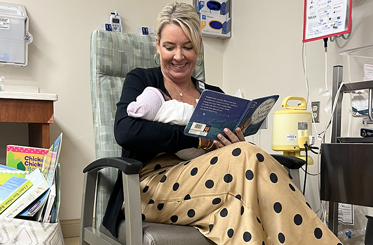 A woman sitting in a chair holds a newborn while reading a book.