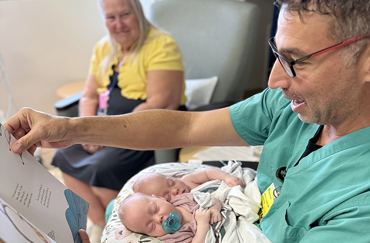 A man in green scrubs holding twin newborns reads a baby book as a woman in background looks on.
