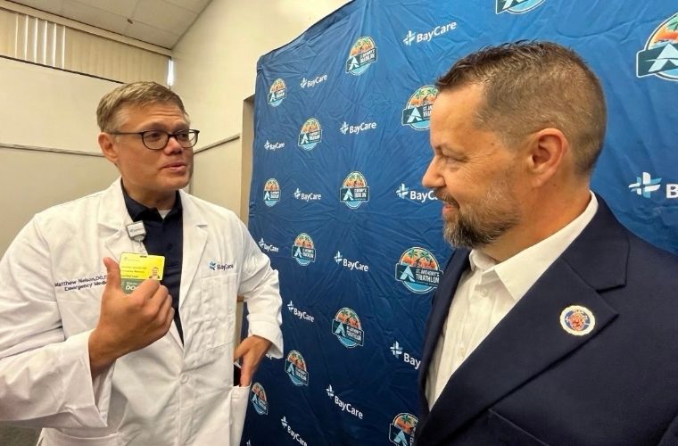 A man in glasses and a doctor's white coat talks with a man wearing sports jacket and a white shirt in front of a blue step and repeat background.