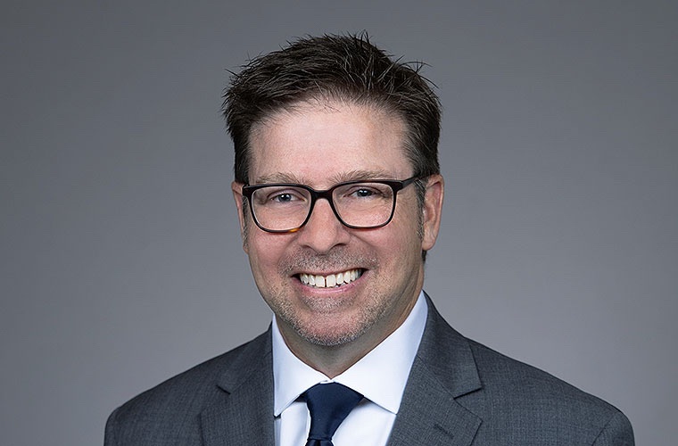 Man with spiky brown hair and brown glasses and wearing a dark gray suit, white shirt and navy-blue tie smiles into the camera.