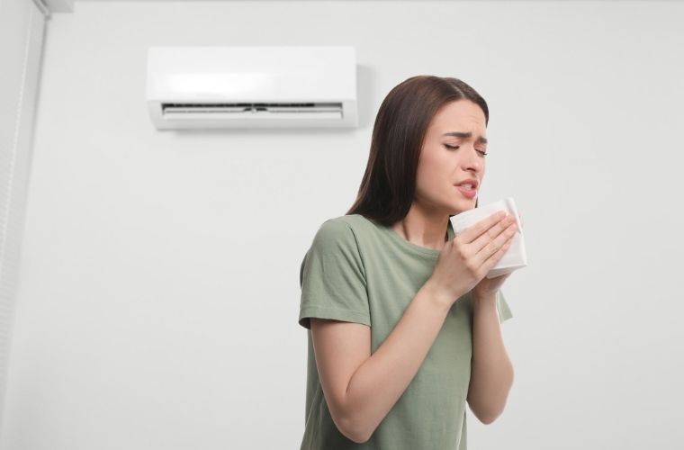 Person standing indoors beneath a wall-mounted air conditioning unit, holding a tissue near their mouth and nose. The individual appears to be experiencing respiratory discomfort, with a plain light-colored wall in the background.