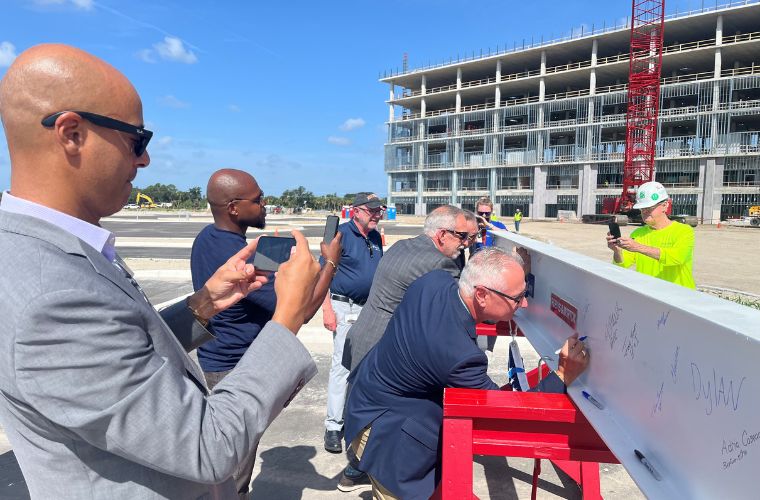 Several BayCare teams members sign a white steel beam at the BayCare Hospital Manatee topping out ceremony while others take photos. The beam rests on red support stands in the foreground, with the multi-story hospital structure and a construction crane visible in the background under a clear blue sky. The image captures a ceremonial construction milestone as the future hospital continues to take shape.