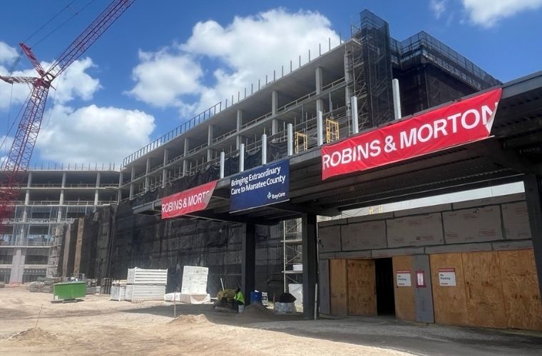 Exterior view of BayCare Hospital Manatee under construction. A multi-story steel-framed hospital structure rises behind an entrance canopy displaying BayCare and Robins & Morton banners. Construction cranes, scaffolding and building materials are visible around the site beneath a bright blue sky with scattered clouds.