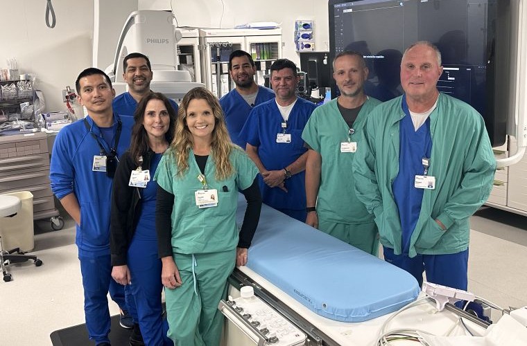A group of eight medical professionals stand in a hospital room. They are gathered around a medical examination table. The room contains advanced medical equipment. The individuals are wearing hospital scrubs and ID badges.