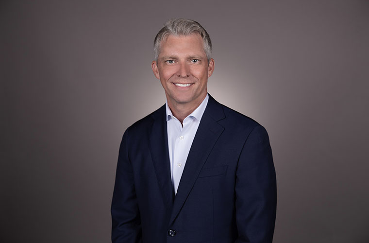 A professional headshot of Patrick Downes wearing a navy suit jacket and white dress shirt against a gray background.