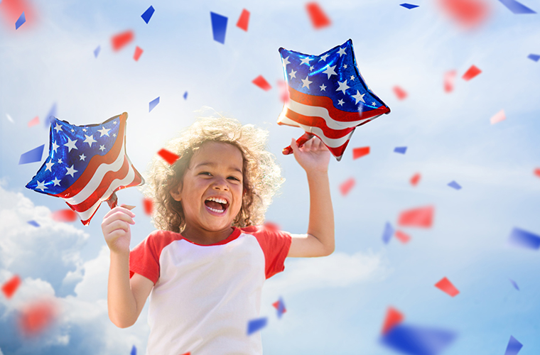 A young child holding star-shaped balloons that are decorated like the American flag. There is red and blue confetti falling in the background.