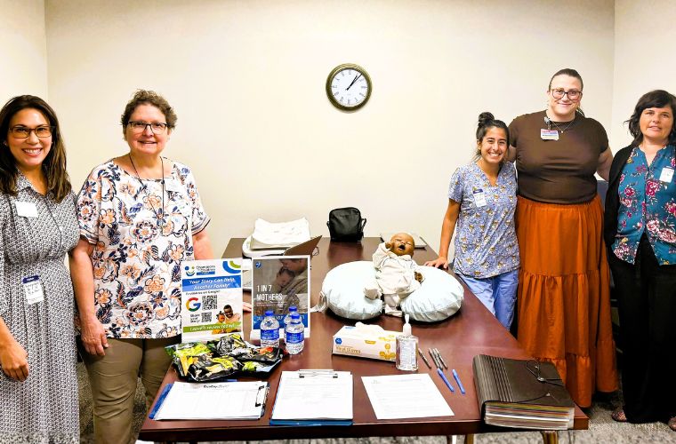 Five representatives from Champions for Children and BayCare stand around a table displaying Baby Café educational materials, supplies and a breastfeeding support demonstration doll at Mease Countryside Hospital.