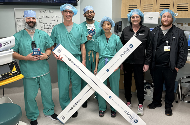 A group of six medical professionals are standing in a hospital room. Five are wearing green surgical scrubs and blue hair covers, while one is in dark clothing. Two individuals are holding large boxes with "UV-CAD" printed on them. Medical equipment and storage cabinets are visible in the background.