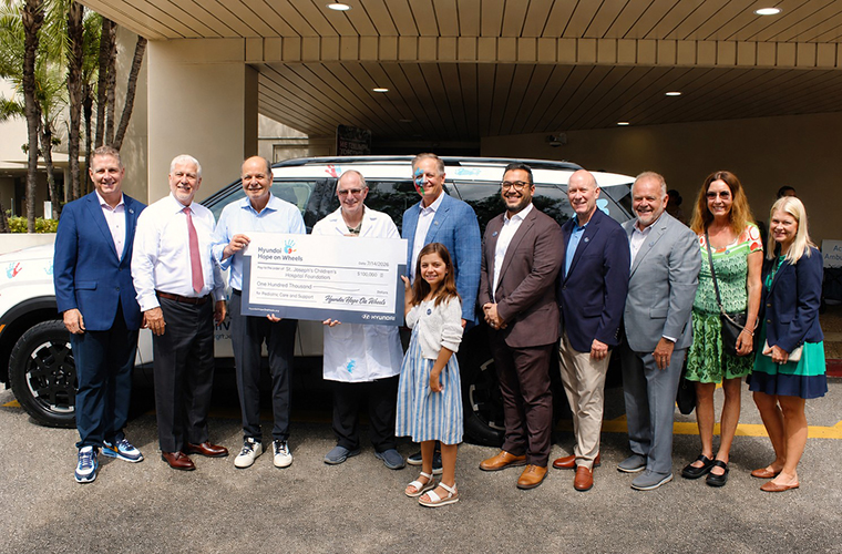 A group of thirteen people standing in front of a building, holding a large ceremonial check. A vehicle is partially visible in the background. The individuals are dressed in formal and semi-formal attire, and they appear to be celebrating or commemorating an event.