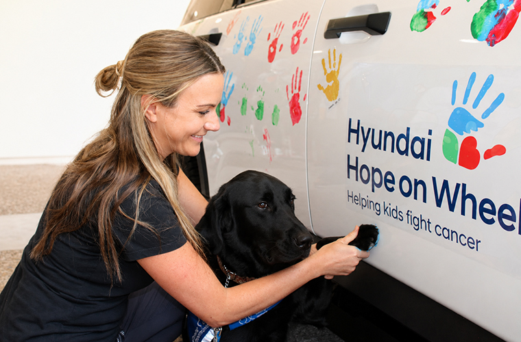 A person gently presses a black dog's paw against the side of a white vehicle. The vehicle is decorated with colorful handprints and features the logo "Hyundai Hope on Wheels" with the slogan "Helping kids fight cancer.
