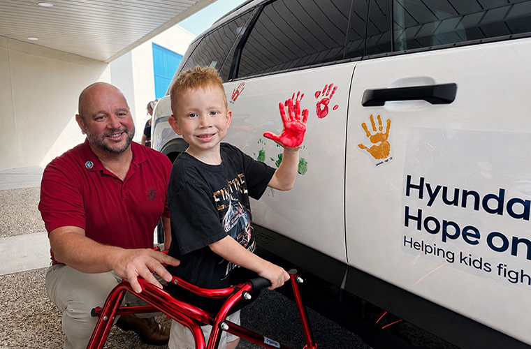 A person and a child are beside a Hyundai car with handprints on it. The child has a red-painted hand and is using a walker. The car has the text "Hyundai Hope on Wheels" on it.