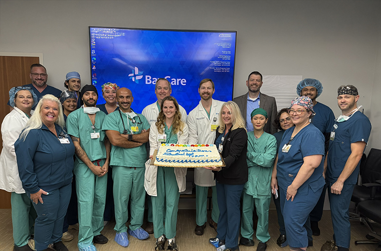 A group of medical professionals wearing scrubs stand in a room with a "BayCare" sign on a screen behind them. They are holding a cake decorated with "Congratulations Surgical Team!" and appear to be celebrating.