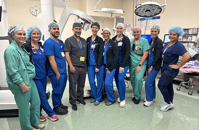 A group of healthcare professionals in scrubs and surgical caps stand smiling in a hospital operating room. The room is equipped with medical machinery and overhead lights.