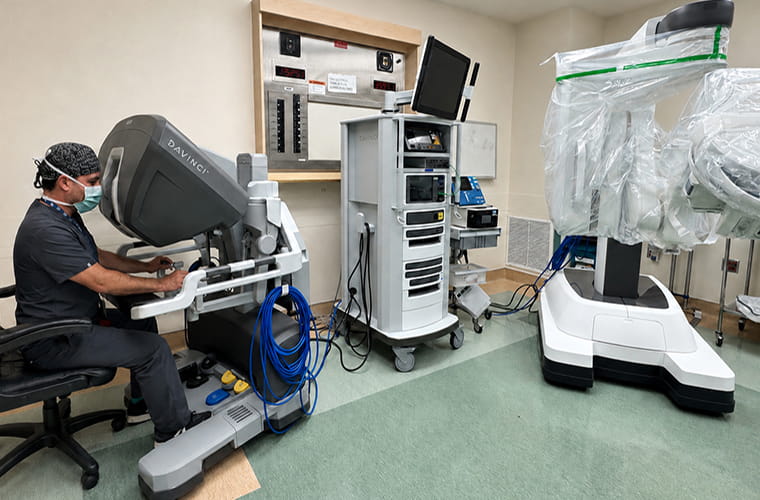 A person operates the a Da Vinci Surgical System in a medical setting. The system includes a console with controls and multiple screens. Other equipment and a robotic unit are positioned nearby. The room has a sterile, clean appearance.
